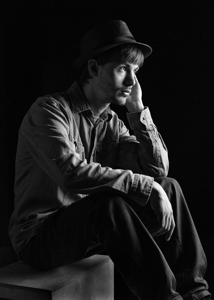 Studio lighting with the male model. Model sitting looking up and wearing a hat. Black and white portrait. Low key portrait. Minimalist lighting setup.
